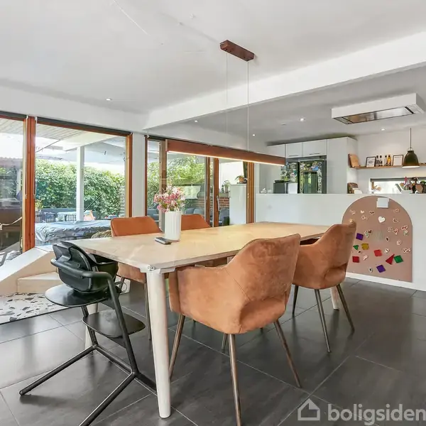 Spisebord står i en moderne spisestue med brune og sorte stole. Bagved er et lyst køkken med indbyggede reoler. Store panoramavinduer åbner op til en terrasse med grønne planter.