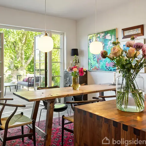 Vase med blomster står på et træbord i en spisestue. Bordet er omringet af stole, og dobbeltdøre åbner ud til en terrasse. Lyse lamper hænger fra loftet.