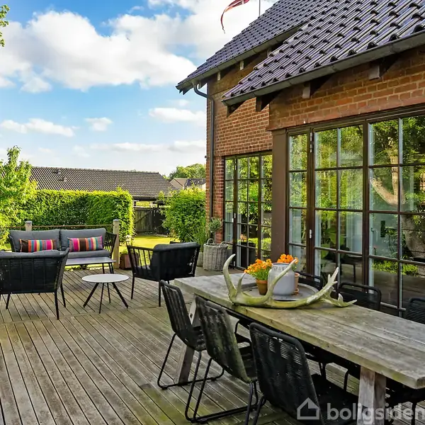 Træterrasse med spisebord og stole, arrangeret til udendørs spisning. Blomsterpotter og en kanne pryder bordet. Baggrund med have, hække og et hus med store vinduer under en blå himmel.