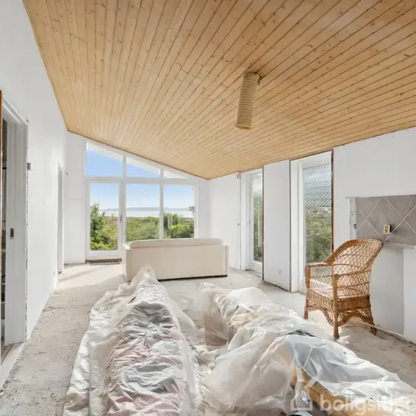 En stue med trægulv og træloft indeholder en hvid sofa, en kurvestol og et vindue med udsigt til grønne omgivelser. Møbler er dækket med plastik for renovering.