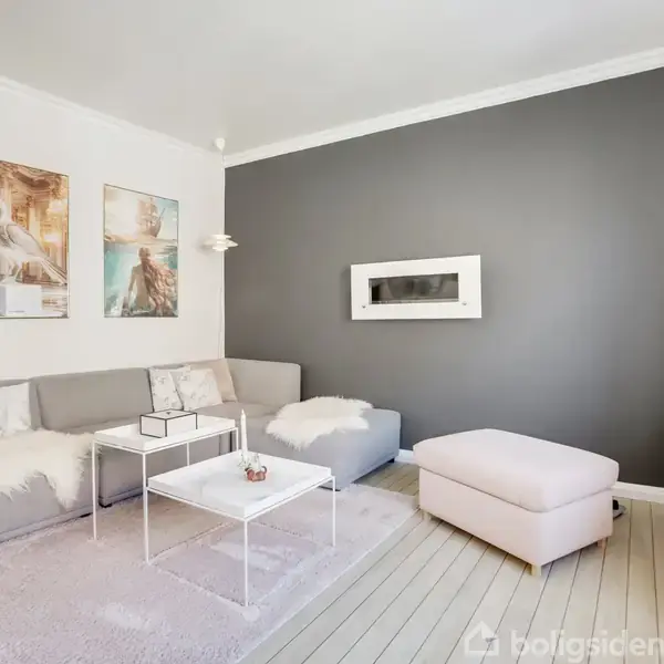 Sofa med hvide tæpper står i en lys stue. Et bord med pynt foran sofaen; billeder hænger på væggen. En reol med lysestager står i hjørnet.