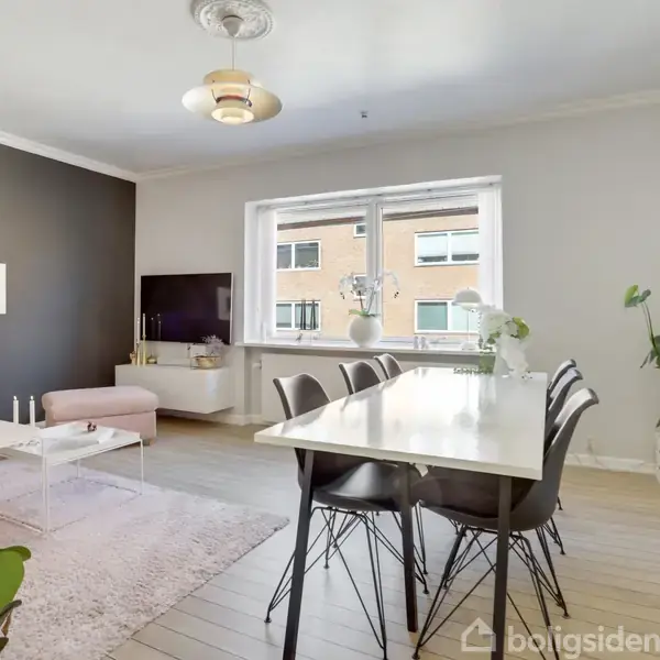Et lyst og moderne indrettet stueområde med spisebord og stole. Vinduer i baggrunden giver naturligt lys. En sofa og TV er placeret ved en mørk accentvæg. Grønne planter tilføjer friskhed.