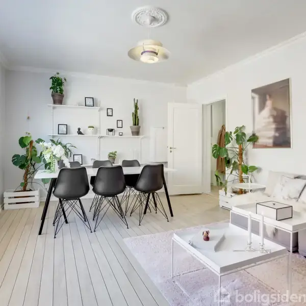 Stue med moderne indretning; en sofa står ved siden af et sofabord, mens en spisebord med sorte stole står nær væggen. Væggene har malerier og hylder med planter.