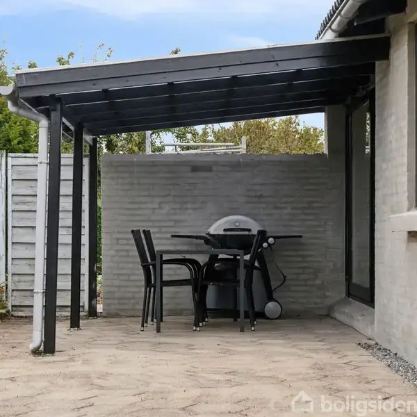 En gasgrill og et bord med tre stole står under en sort overdækning på en flisebelagt terrasse ved siden af et hus. Hegn og grøntsager omgiver terrassen.