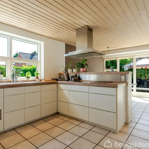 Et moderne køkken med hvide skabe og træbordplader. Kogezonen har en emhætte over. Store vinduer bader rummet i naturligt lys, og udenfor ses en terrasse med et bord og parasol.