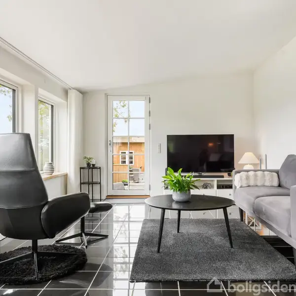 Stue med grå sofa og sort lænestol placeret på sort flisegulv. Tv’et står foran en terrassedør, mens et sofabord med en plante står centralt. Lyse vægge og vinduer.