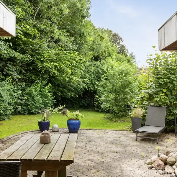 Træbord med dekorativ figur står på en terrasse omgivet af grønne planter og buske. To loungestole står i nærheden, og området har en hyggelig, afslappet atmosfære.