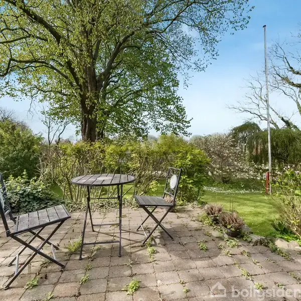 Et rødt murstenshus står stille ved en grusvej, omgivet af grønne træer og en velplejet have. Huset har flere vinduer og et skifertag, hvilket tilføjer charme til omgivelserne.