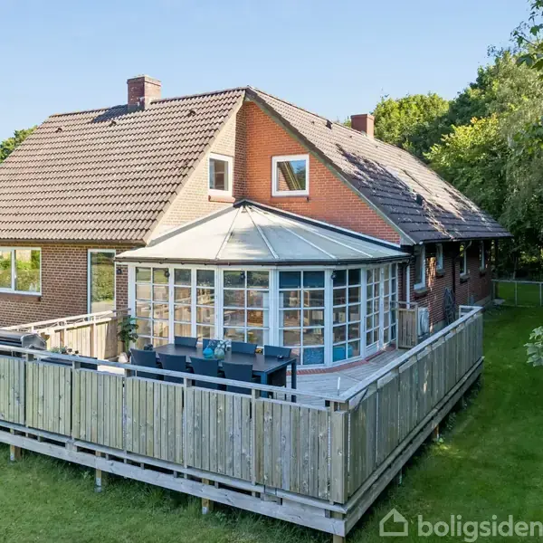Et rødt murstenshus med en stor udestue og træterrasse. Terrassen har et spisebord og stole. Huset er omgivet af en grøn have og træer i baggrunden.