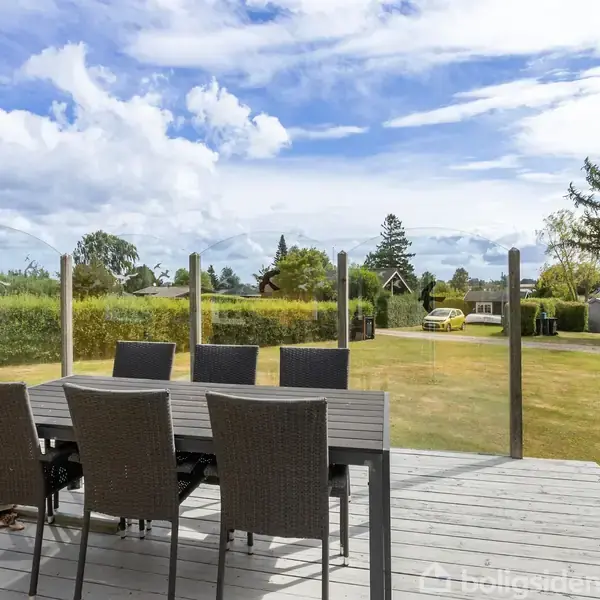 En udendørs terrasse med et spisebord og seks stole står på et trædæk. Baggrunden viser en velplejet have og en klar blå himmel med skyer.