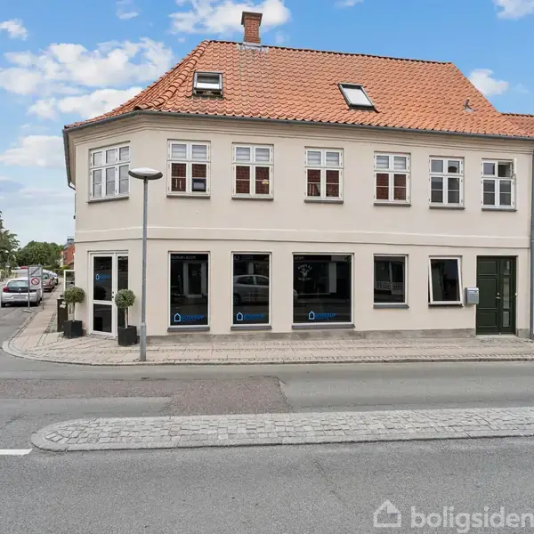 En hvid to-etagers bygning med rødt tegltag står på en gadehjørne. Flere vinduer pryder facaden. Bygningen er omkranset af en stille vej og en fortov.