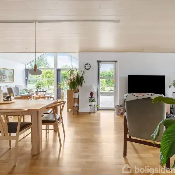 Stue med spisebord og stole i træ, placeret ved store vinduer, der giver udsigt til en terrasse. Der er planter, en vægmonteret fladskærm og naturligt lys, der fylder rummet.