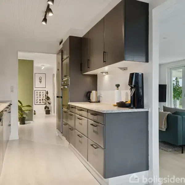 Køkken med mørke skabe og hvide bordplader, hvorpå en kaffemaskine står. I baggrunden ses stue med grøn sofa og lyse vægge. Plant og kunstværk pryder gangen.