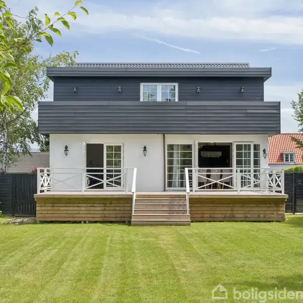 Et moderne hus med en to-etagers træterrasse, omringet af en velholdt græsplæne. Ved siden af ses træer og et rødt tag i baggrunden.