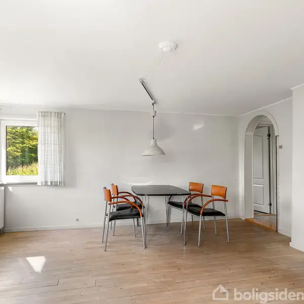 Et spisebord med fire stole står i et lyst rum med trægulv. En stor lampe hænger over bordet. Vinduet til venstre giver udsigt til grønne træer.