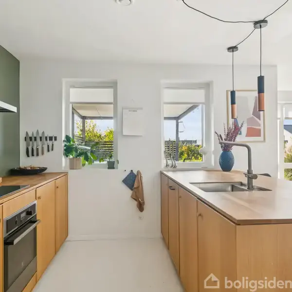 Et moderne køkken med lyse træskabe og hvide vægge har en indbygget ovn til venstre. Knive hænger på væggen, og vaser med blomster står på en køkkenø. Gennem vinduer ses en terrasse og have.