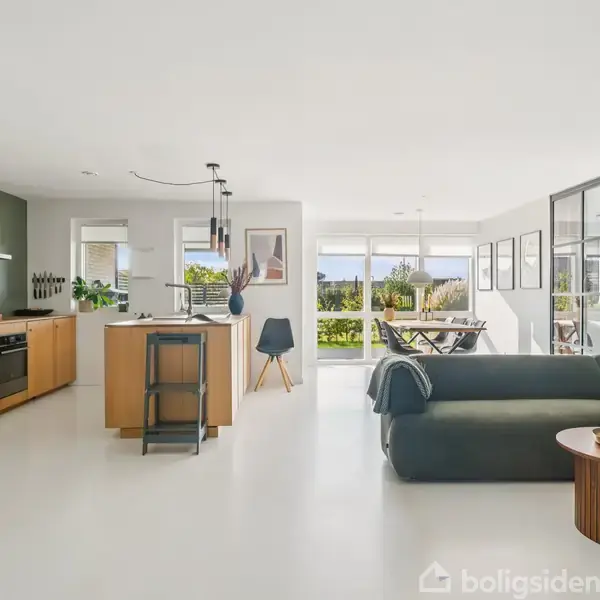 Et moderne, åbent køkkenalrum med et grønt sofaarrangement. Køkkenet indeholder hvidevarer og en central ø. Store vinduer slipper lys ind og tilbyder udsigt til en grøn have.