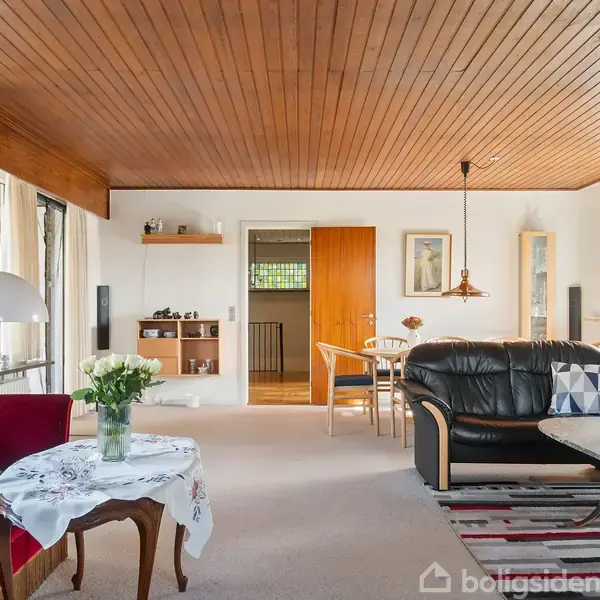 Rød sofa og sort læderlænestol står i en stue med træloft og store vinduer, der giver masser af lys. Et spisebord med stole ses i baggrunden.