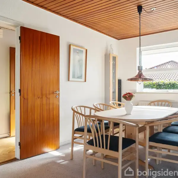 Et spisebord med seks stole står i et lyst rum. En vase med blomster står på bordet. En dør er åben mod en anden del af huset.