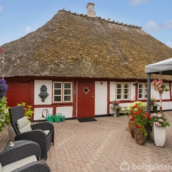 Et gammeldags bindingsværkshus med stråtag står i en brostensbelagt gård. Omkring huset er der udendørs møbler og blomsterkrukker. Himmel er blå med få skyer.