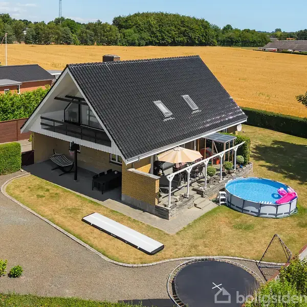 Huset med sort tag står i en indhegnet have med trampolin og swimmingpool. På terrassen er der en parasol, omgivet af et landligt landskab med gule kornmarker i baggrunden.