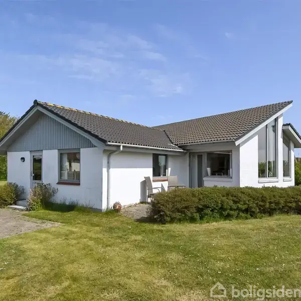Hvidt, moderne hus med store vinduer, står på en grøn græsplæne. Omkranset af buske, beliggende i et naturskønt område under en klar blå himmel.