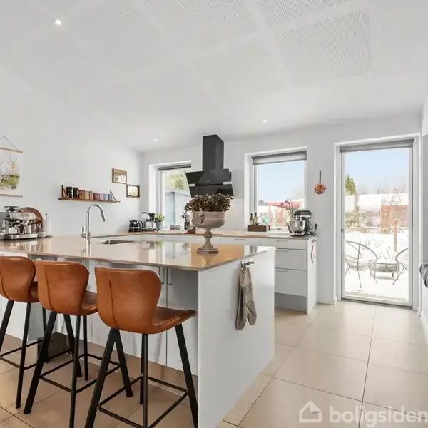 Køkkenø med tre brune barstole sidder foran den. Lysindfald fra store vinduer på højre side skaber en lys atmosfære. Moderne indretning med hvidt interiør og beige gulvfliser.