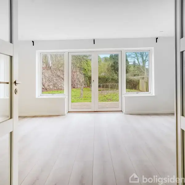 Dobbelt glasdør åbner ind til et lyst rum med trægulv, store vinduer og udsigt til en grøn have med stakit.