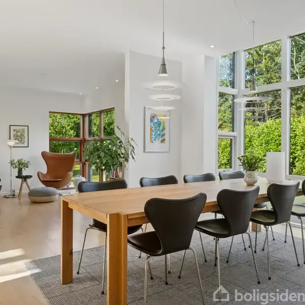 Et spisebord med sorte stole står i et lyst rum med store vinduer og en udsigt til en grøn have. Til venstre ses en lænestol og flere potteplanter.