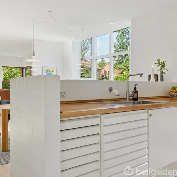 Hvid køkkenø med trærækkebordplade rummer en vask og planter. I baggrunden ses et lyst spiseområde med store vinduer, der giver udsigt til grønne træer udenfor.