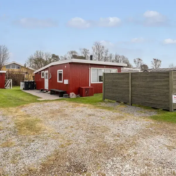 Rødt træhus står stille omgivet af en grøn græsplæne og grusvej. Til højre ses en træhegn med et skilt "Mægler", omgivet af flere buske og træer i baggrunden.