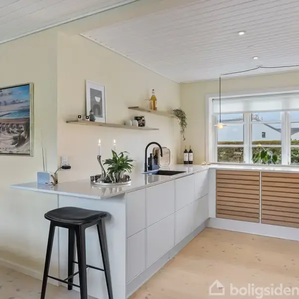 Moderne køkken med hvidt interiør. Barstol ved en køkkenø under sort vandhane på bordpladen. Store vinduer bagved lukker dagslys ind. Indrammet billede og planter tilføjer dekoration.