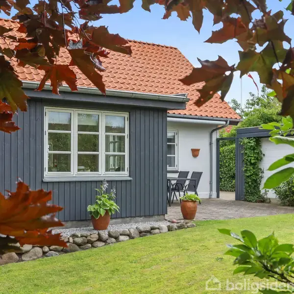 Et hus med rødt tegltag omgivet af en grøn have. Der står en plante i en krukke ved vinduet, og en terrasse med havemøbler ses i baggrunden.