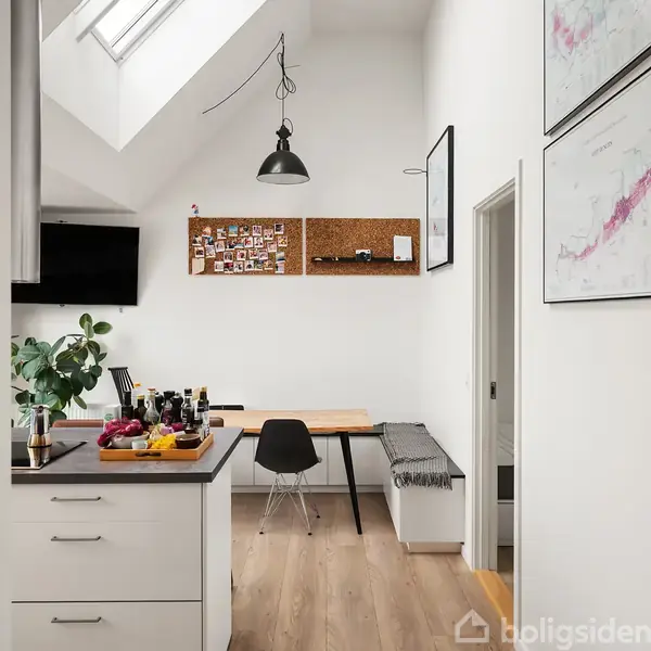 Et moderne køkken med et hvidt interiør. Et spisebord med en sort stol og bænk står mod væggen. En opslagstavle og planter skaber en hyggelig atmosfære.