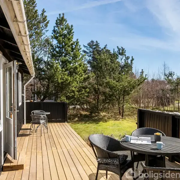 Terrassebord med stole står på en træterrasse, omgivet af en have med græs og træer. Vejret er solrigt, og der er to bærende kopper på bordet.