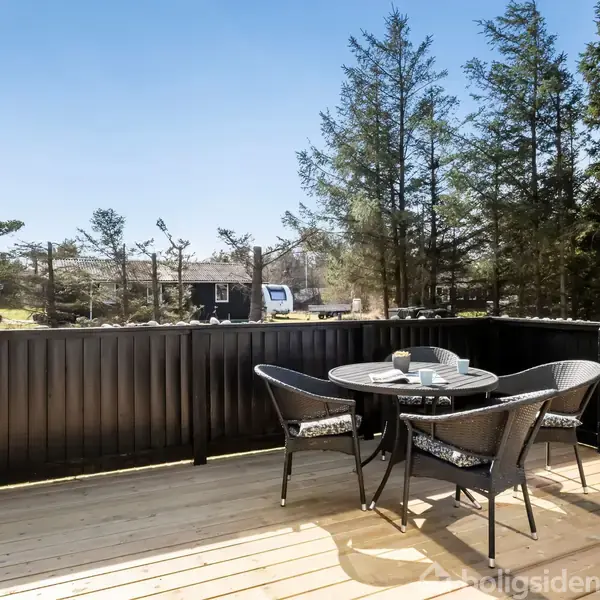 Rundt havebord med tre stole står på terrasse i træ omgivet af sort stakit. Bordet er dækket med tallerkener i solbeskinnet have med høje, grønne træer i baggrunden.