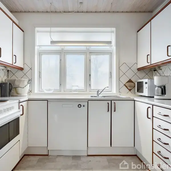 Et rummeligt køkken med hvide skabe og sorte håndtag. En vask er placeret under et vindue. Omgivet af køkkenapparater som ovn, mikroovn og kaffemaskine.