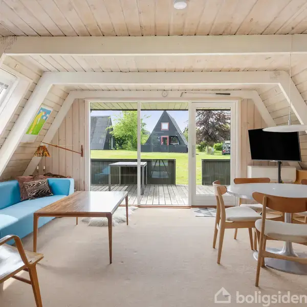 Stue med blå sofa og træmøbler, der vender mod en stor glasskydedør, der fører til en terrasse. Udenfor ses en have med en triangulær bygning.
