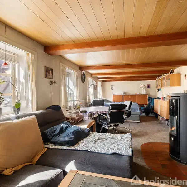 Stue med grå sofa og bord, hvor lys strømmer ind gennem vinduer. En brændeovn står i nærheden, mens hylder og møbler fylder rummet, der har et træloft med synlige bjælker.