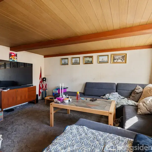 Stue med hjørnesofa dækket af tæpper og puder, sofabord med lysestager, fladskærms-tv på skænk, rødt flag, væg billeder. Loftet er træbelagt, vindue med lysindfald.