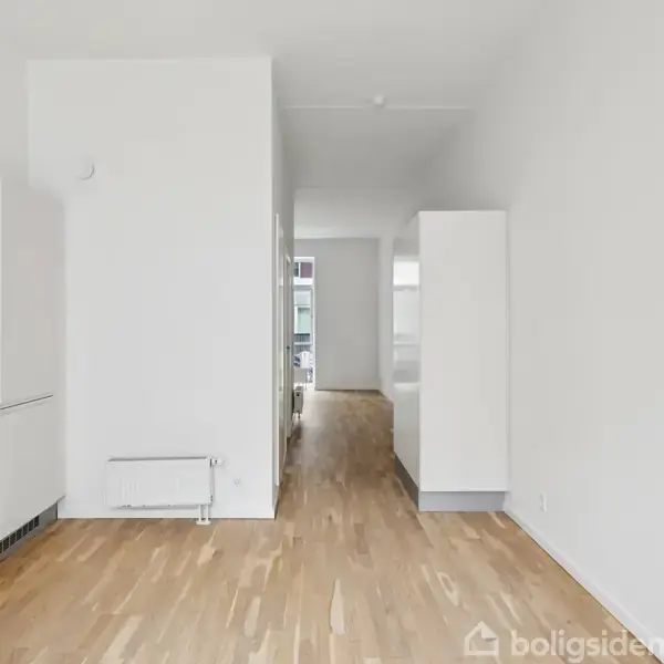 Et hvidt, minimalistisk køkken med trægulv fører til en lys gang. Skabe og radiator ses til venstre, og et vindue anes for enden af gangen.