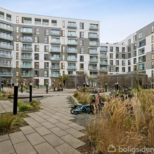 Lejlighedskompleks med flere etager og moderne altaner omgiver en indre gård. Et bredt flisebelagt gangareal løber gennem et område med beplantning og cykler parkeret ved siden af stien.