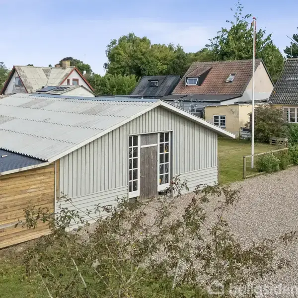En grå bygning med store vinduer står stille på en grusbelagt gårdsplads, omgivet af huse og grønne træer under en blå himmel.