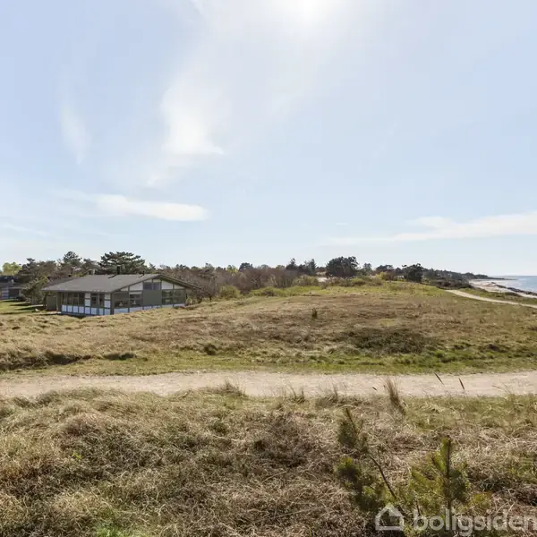 Et hus står stille omgivet af en frodig græsslette nær kysten. En sti snor sig gennem landskabet mod havet under en lysende blå himmel.