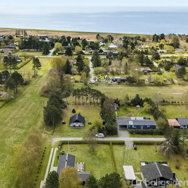 Huse ligger spredt langs grønne marker og træer med udsigt over en kystlinje i horisonten. Små veje forbinder boligerne i dette landlige område.