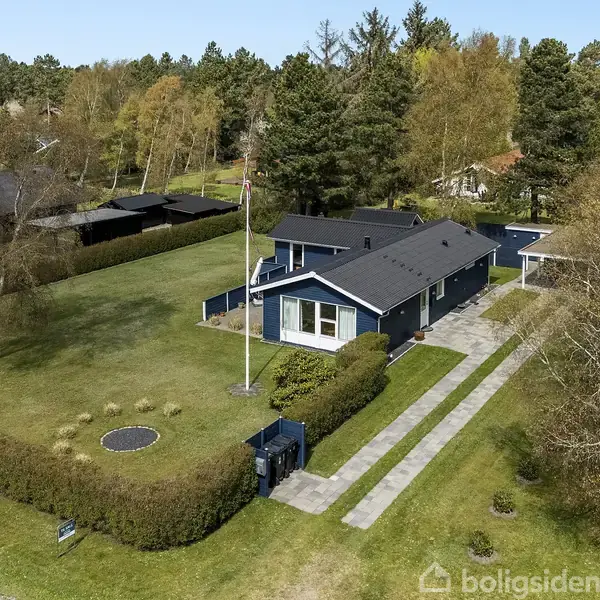 Et blåt enfamiliehus med flagstang står i en velplejet have omkranset af hække. En sti leder til huset, omgivet af træer. Trampolin i græsset ved siden af indgangen.