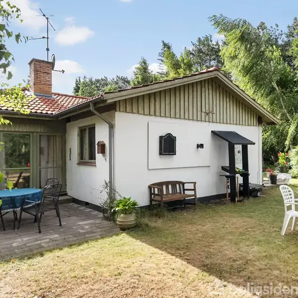 Et hvidt hus med rødt tegltag og terrasse i haven. Terrassen har et rundt bord med blå dug og sorte stole. En grill er placeret ved en væg.