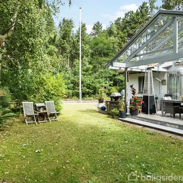 En glaspavilion med terrasse, omgivet af frodig have og skov. Der er udemøbler på terrassen og en lille ildsted i haven. Græsplænen forbinder forskellige siddepladser.
