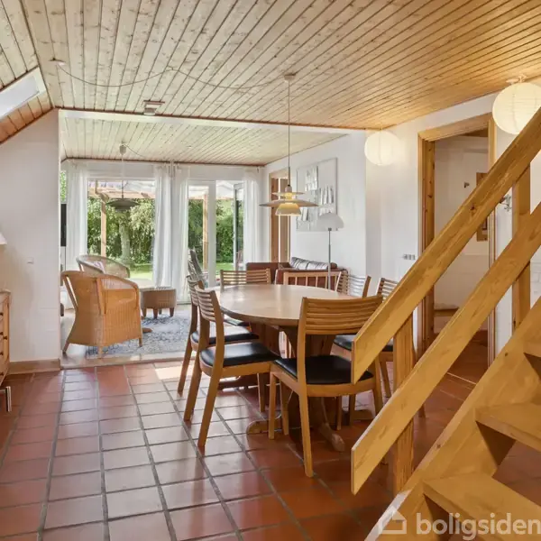 Spisestuen har et rundt træbord med stole. Gerne udsigt gennem glasdøre til haven udenfor. Træloft og terracotta gulvfliser dominerer, mens en trappe fører opad.