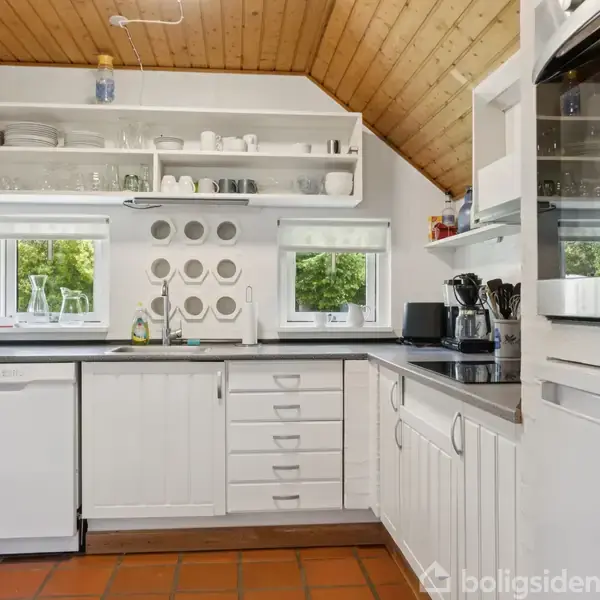 Et lyst køkken med hvide skabe og åben hylde med service. Vask med to vinduer over, opvaskemaskine, ovn, og køkkenredskaber på højre side. Træloft og terracotta-flisegulv.